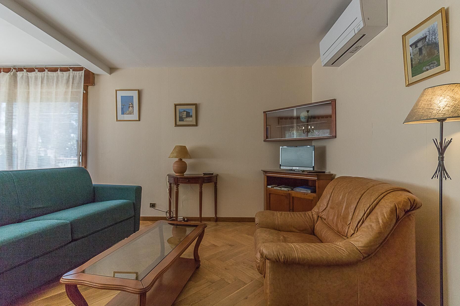 Ein Wohnzimmer mit einem türkisfarbenen Sofa, einem braunen Ledersessel, einem Glastisch, einem Fernseher auf einem Holzständer, einer Lampe und gerahmten Bildern an der Wand.