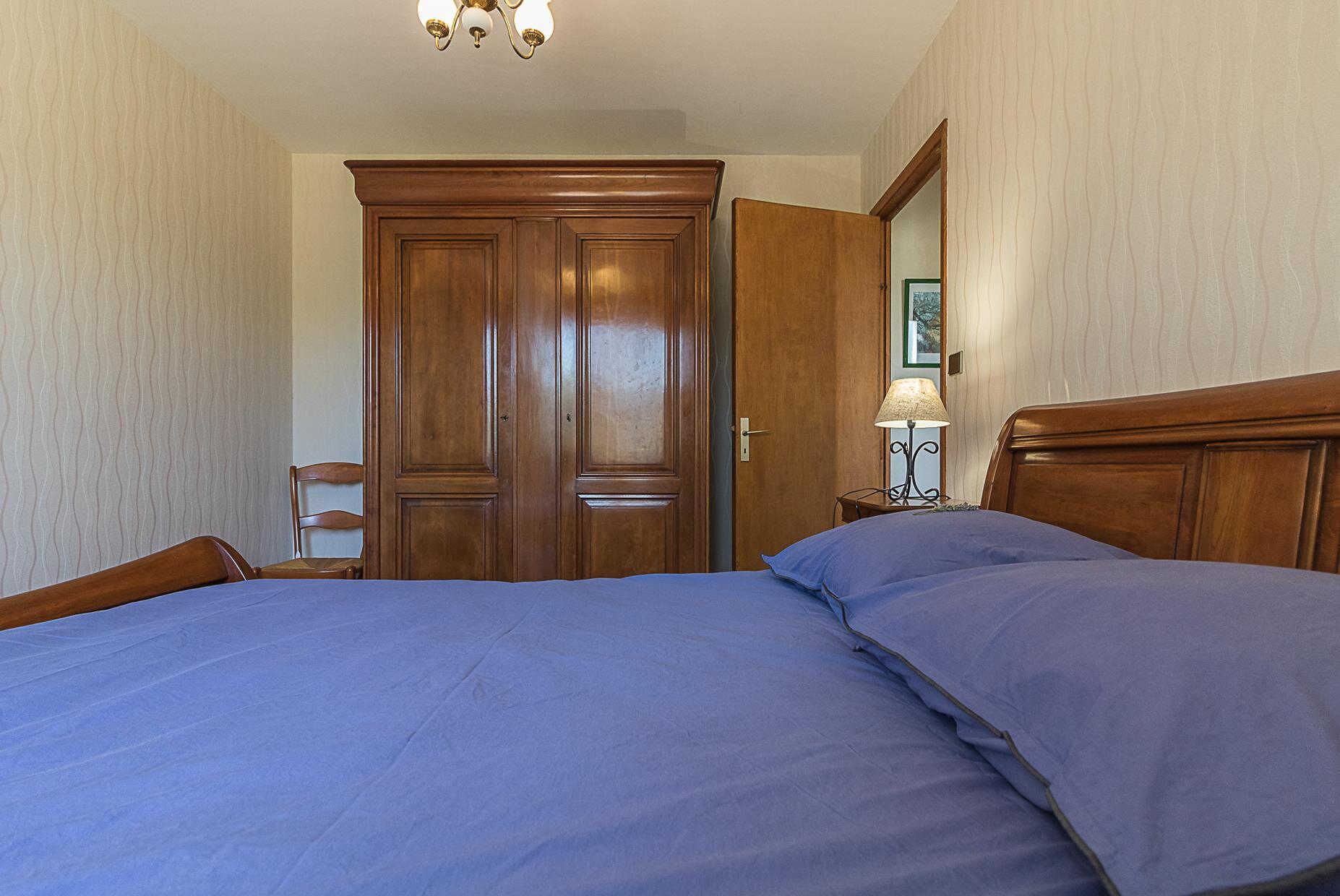 A bedroom with a double bed, wooden wardrobe, and a nightstand with a lamp.