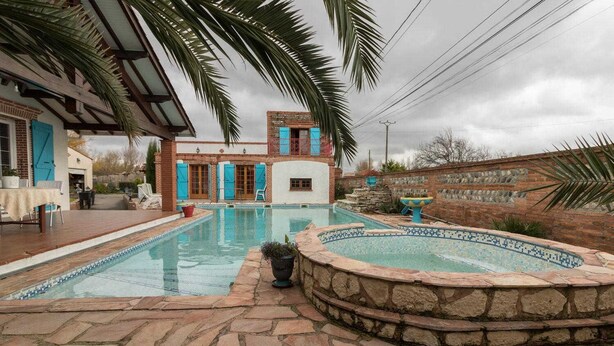 Ein Schwimmbecken mit einer Steindeck, eine kleine Whirlwanne und ein Gebäude mit blauen Fensterläden.