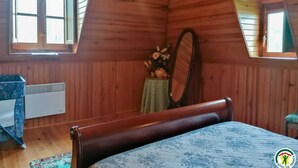 A bedroom with a sloped ceiling, wooden walls, a bed with a patterned bedspread, a small table with flowers, a mirror, and a window with shutters. - Chez Marthe - Melles (Melles)