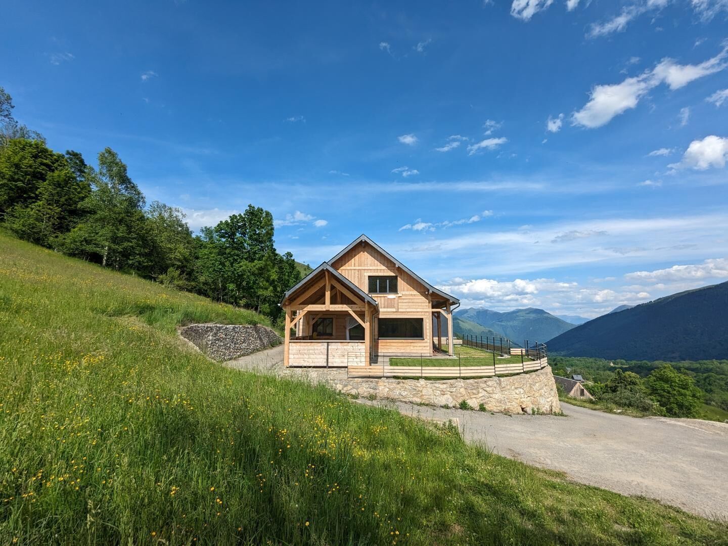 A wooden cabin with a sloped roof, situated on a grassy hill with a stone wall and a paved path leading to it.