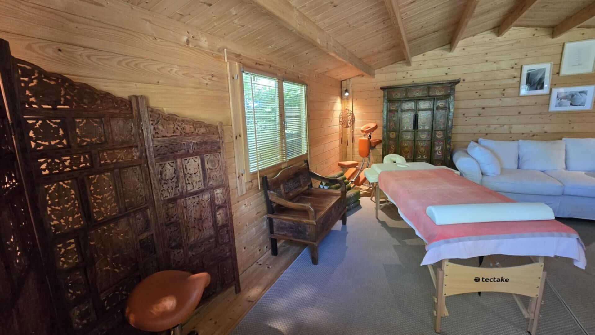 Een houten hutje met een bed, een bank, een massagetafel en een massagestoel.