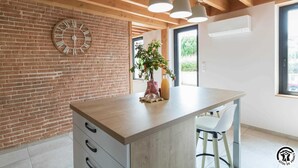 Une cuisine moderne avec un plan de travail en bois, un mur en briques, une grande horloge et une fenêtre donnant sur un paysage verdoyant.