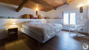 Ein Schlafzimmer mit Bett, Nachttisch, Lampe, Stuhl, Fenster und Holzbalkendecke.