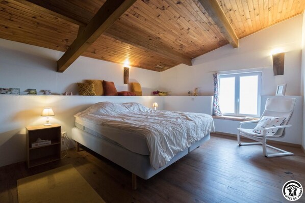 Un dormitorio con cama, mesita de noche, lámpara, silla, ventana y vigas de madera en el techo.