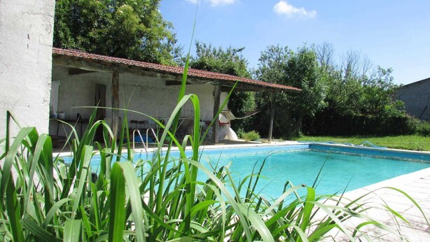 Ein Schwimmbecken mit Fliesen umgeben von Grünflächen und einem Gebäude mit rotem Ziegeldach.