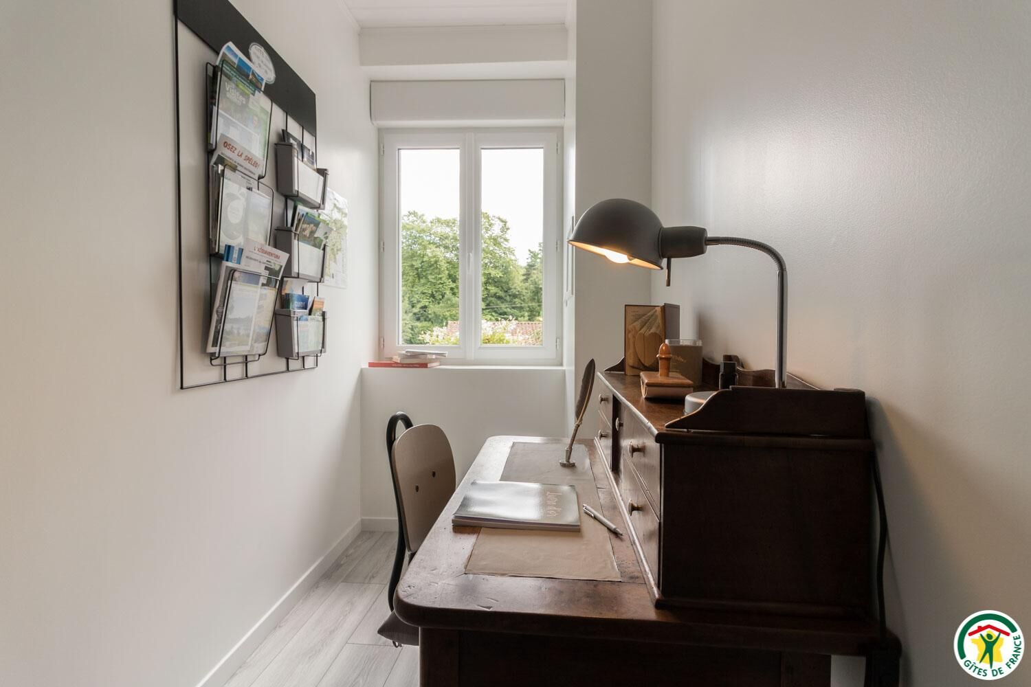 Una habitación con un escritorio de madera, una silla, una lámpara, un ventanal con vistas a follaje, y un tablón de anuncios con papeles y fotografías.