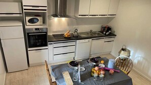 Cocina compacta con armarios blancos, estufa, microondas y fregadero. Hay una mesa con mantel oscuro, varios utensilios de cocina y una cesta de frutas.