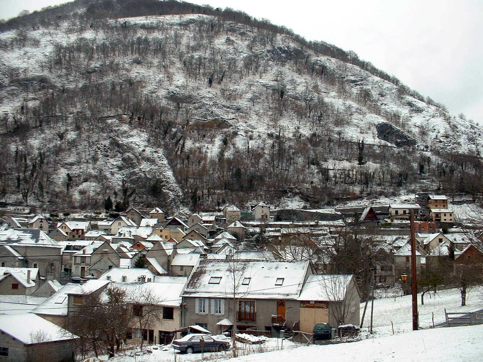 Un village enneigé, avec des maisons regroupées autour d’une place centrale, une église et une montagne en arrière-plan.