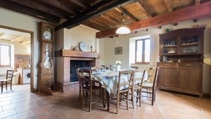 A rustic kitchen with a dining table set for six, surrounded by wooden chairs. A brick fireplace, a clock, and a wooden cabinet with glassware are visible. - Panifous - Montbrun-Bocage (Montbrun-Bocage)