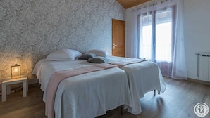 Een slaapkamer met twee bedden, behang met een patroon, een houten plafond, een nachtkastje met een lamp en een raam met dunne gordijnen.
