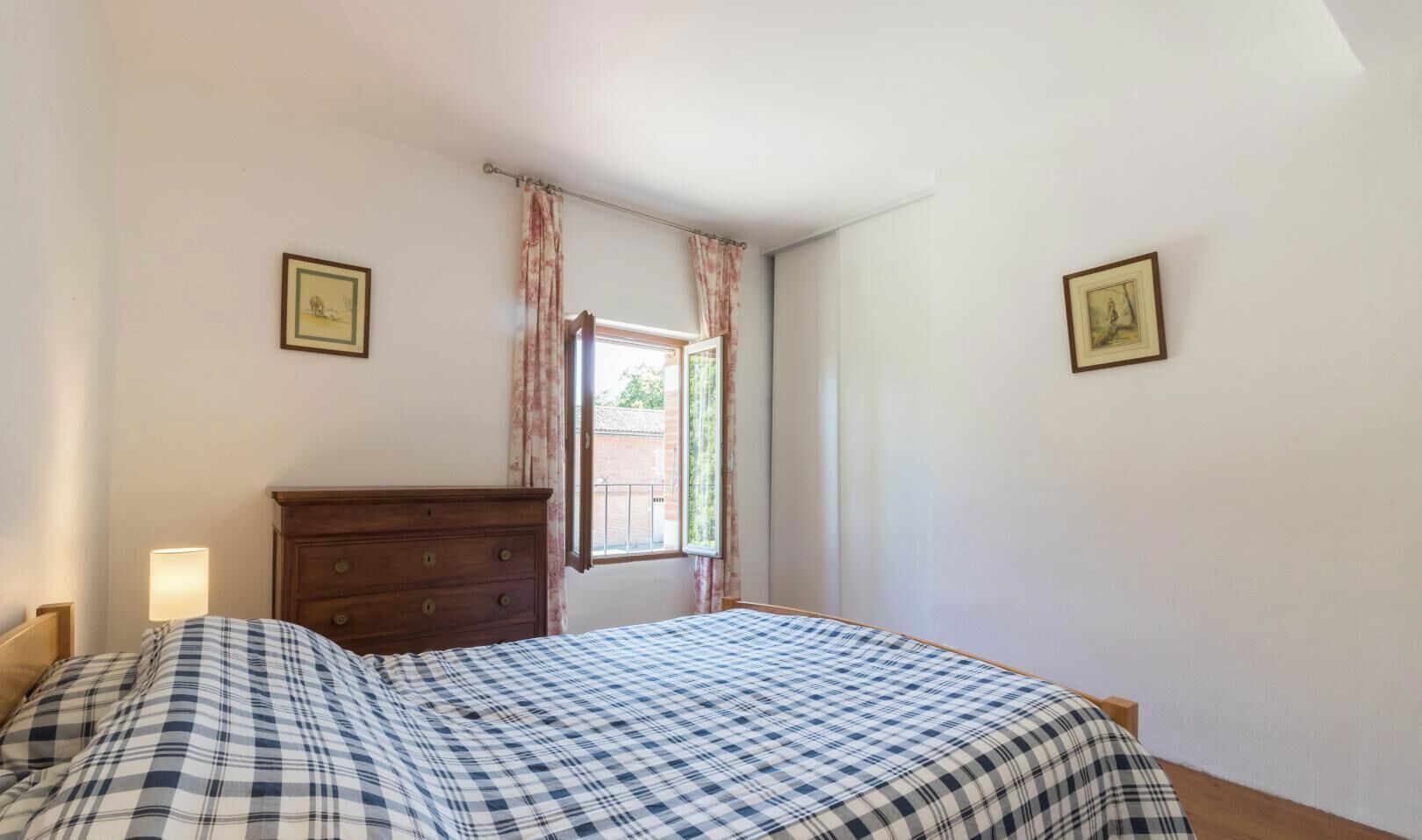 Un dormitorio con cama, cómoda de madera, ventana con cortinas y dos cuadros en las paredes.