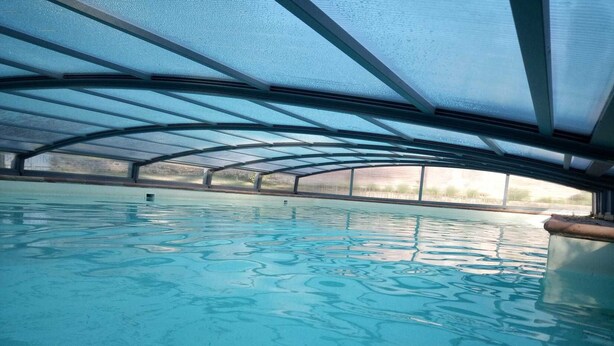 An indoor swimming pool with a clear blue water surface, surrounded by a covered area with a metal roof structure.