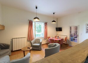 Een woonkamer met een bank, fauteuils, een eettafel met stoelen, een aan de muur bevestigde televisie en een raam met gordijnen.
