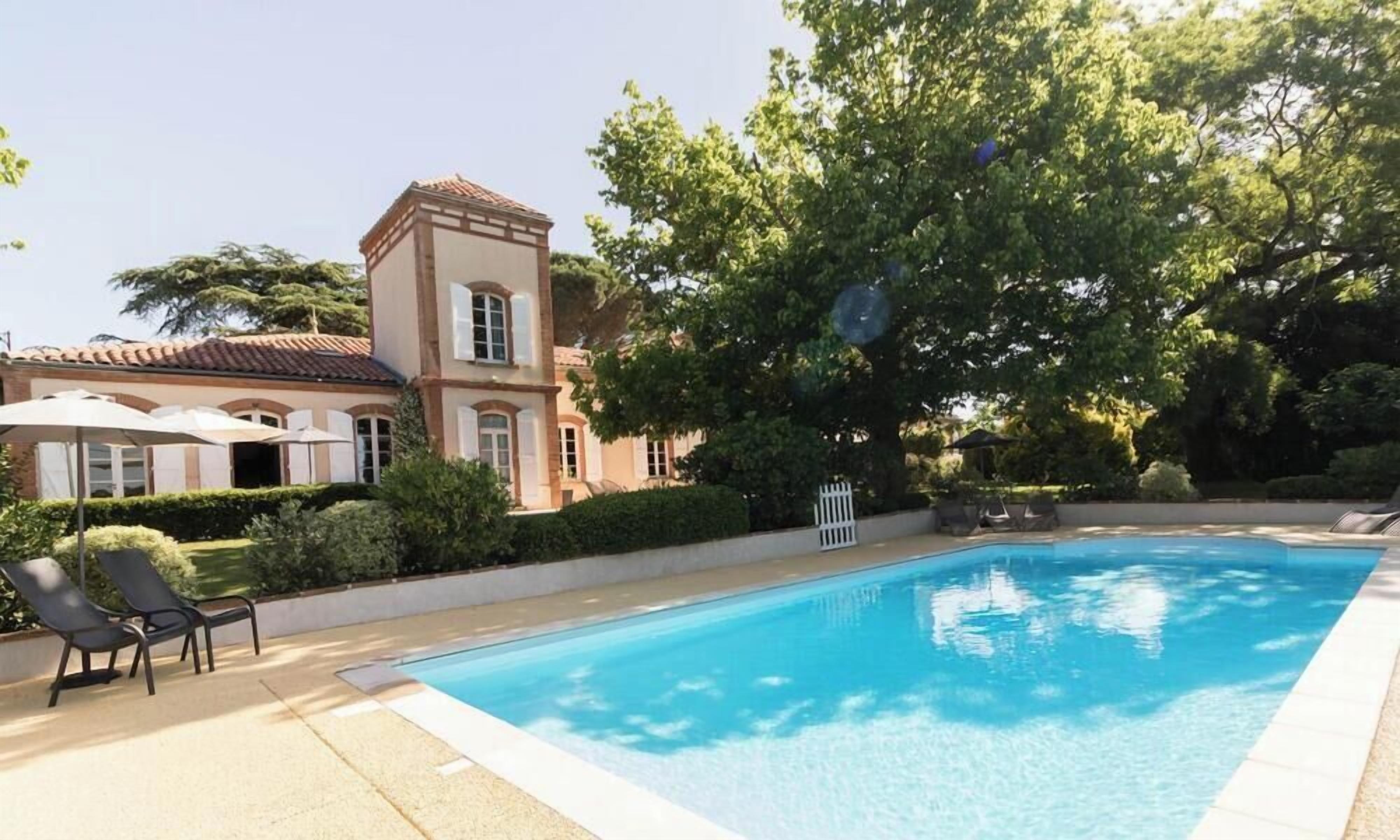 Piscina com espreguiçadeiras, um edifício com telhado em azulejos e vegetação ao redor.