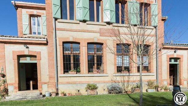 Un edificio de dos plantas con fachada de ladrillo, contraventanas verdes y numerosas ventanas.