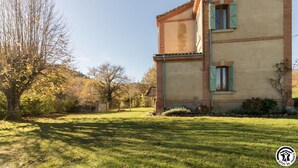 Una casa de dos pisos con fachada de ladrillo y contraventanas verdes, rodeada por un patio cubierto de césped y árboles.