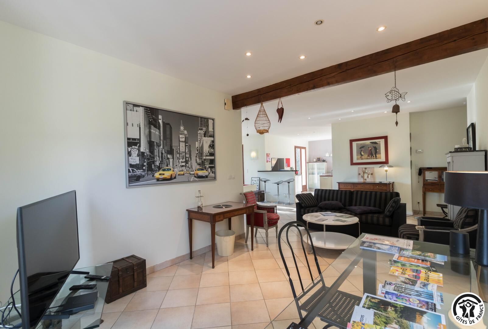 Un salón con un cuadro grande, un televisor, una mesa de comedor con revistas y una cocina al fondo.