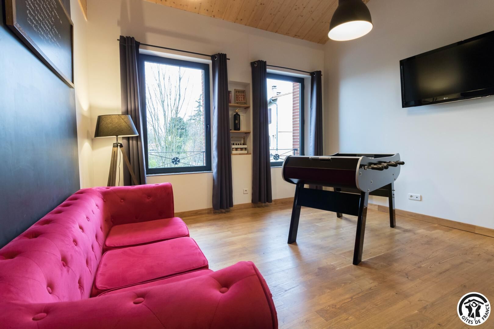 Ein Zimmer mit einem roten Sofa, einem Kicker, einem Fernseher und großen Fenstern mit Vorhängen.
