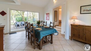 Un coin repas avec une table dressée pour quatre personnes, des chaises en bois, une nappe de couleur turquoise et une vue sur l’extérieur grâce à une porte vitrée.