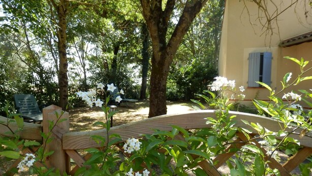 Une terrasse en bois entourée d’une végétation luxuriante et de fleurs blanches, avec un arbre et un bâtiment en arrière-plan.