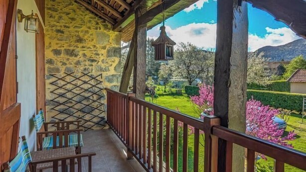 Un'area salotto esterna riparata, con pareti in pietra, una ringhiera in legno e vista su un giardino e montagne.