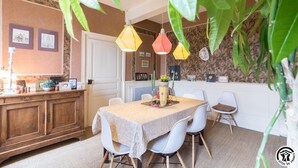 Ein Essbereich mit Tisch, gedeckt für vier Personen, umgeben von weißen Stühlen. Über dem Tisch hängen Pendelleuchten, und links steht eine Holzkabine mit Spiegel und Dekoration.
