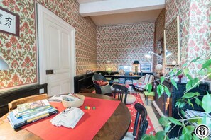 Uma sala de estar acolhedora com papel de parede estampado, uma área de jantar com livros e toalha de mesa vermelha, e uma lareira.