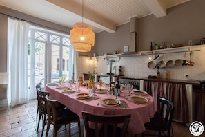 Uma área de refeições com uma mesa redonda posta para seis pessoas, um lustre pendurado e uma cozinha ao fundo.