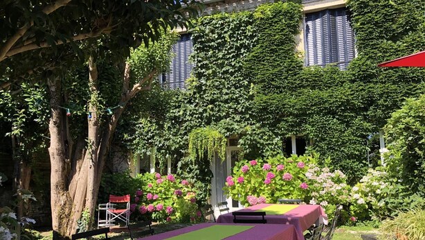 Ein Garten mit Tischen, die mit violetten Tischdecken bedeckt sind, umgeben von üppigem Grün und blühenden Pflanzen.
