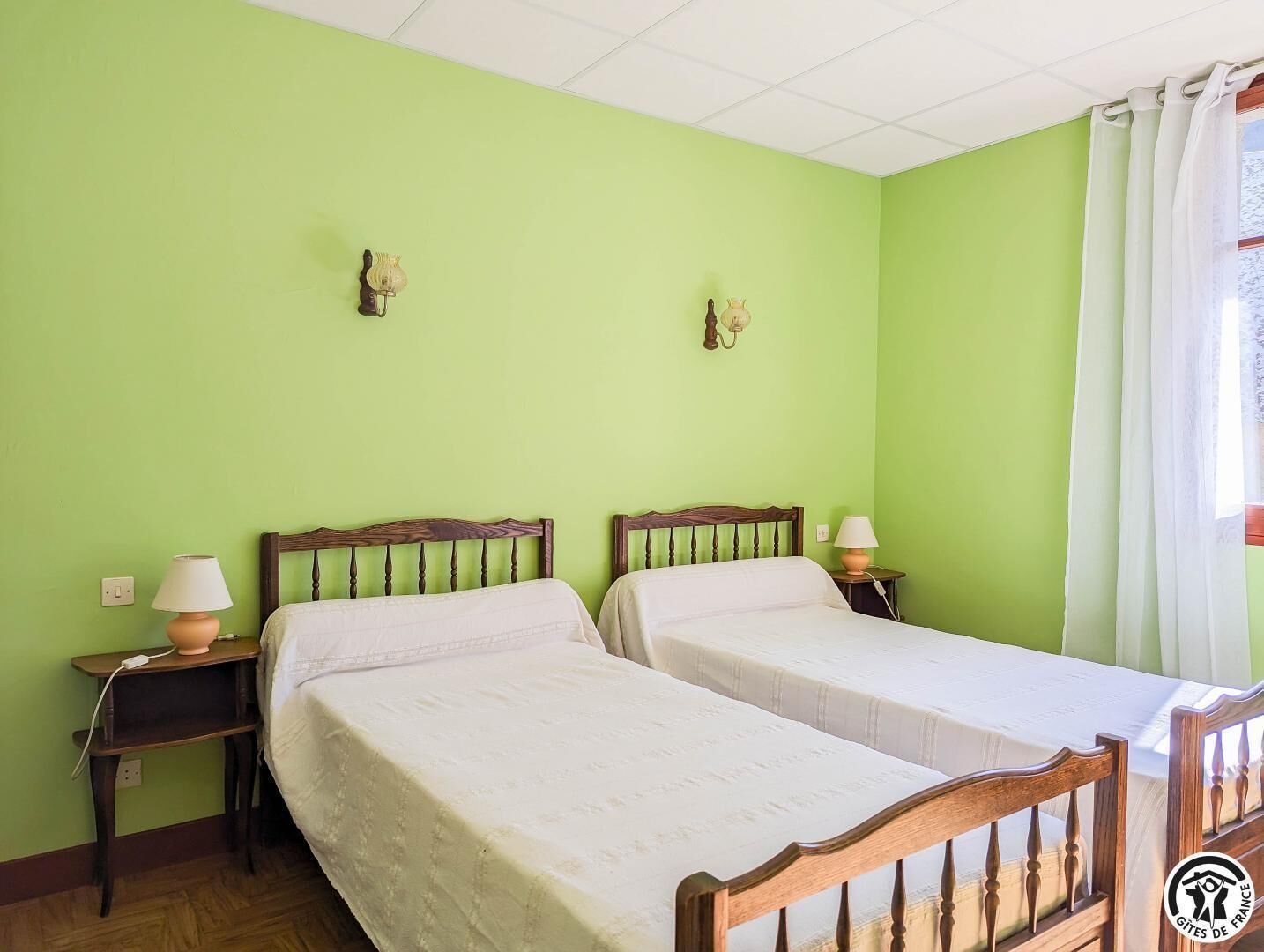 Une chambre avec deux lits simples, chacun doté d’une tête de lit en bois et d’une table de chevet avec une lampe. Les murs sont peints en vert et il y a une fenêtre avec des rideaux blancs.
