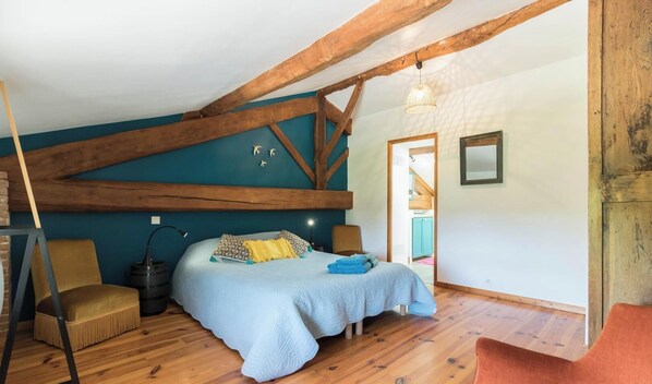 Ein Schlafzimmer mit schrägverdachter Decke, Holzbalken, einem Bett mit blauer Bettdecke, einem Sessel und einer Pendelleuchte.