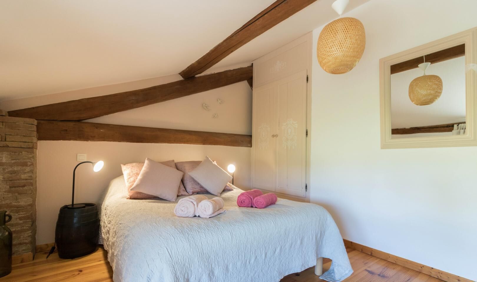A bedroom with a bed, pillows, a bedside lamp, a hanging light, a mirror, and a wooden beam ceiling.