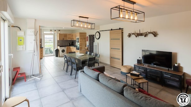 En moderne lejlighed med spiseplads, køkken og opholdsstue. Der er et fjernsyn, en sofa og et spisebord med stole.