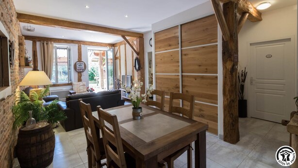 L’intérieur est de style rustique, avec une table à manger en bois, des chaises et un canapé. Un grand tronc d’arbre est mis en valeur, et on peut voir une porte et une fenêtre.
