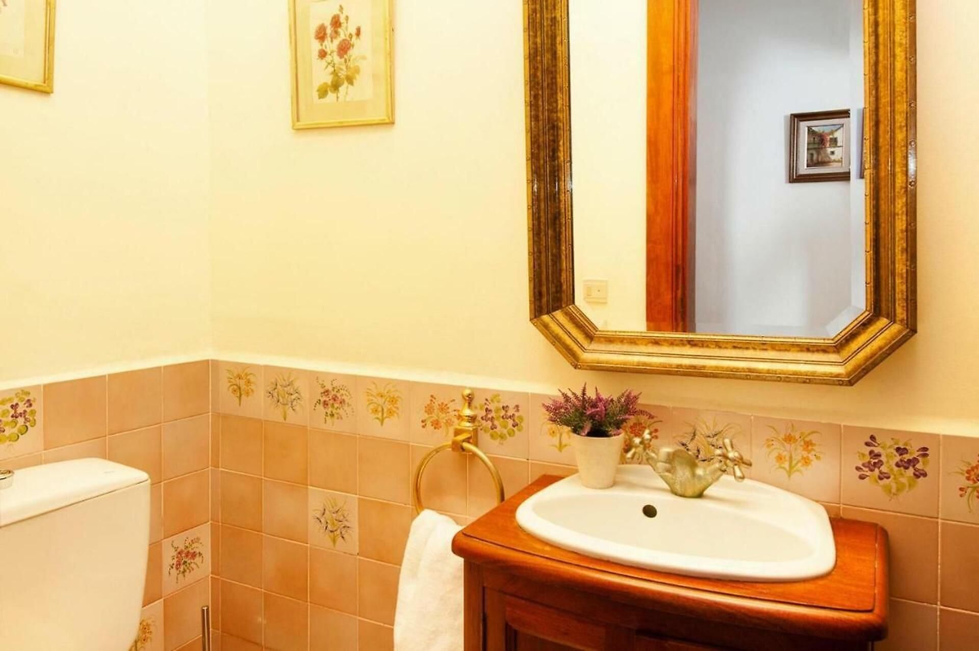 A bathroom with a white sink, a wooden vanity, a gold-framed mirror, and floral tiles on the walls.