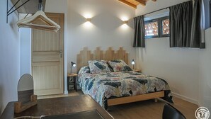 A bedroom with a wooden headboard, a bed with a floral bedspread, a desk with a chair, a hanging clothes rack, and a window with curtains.