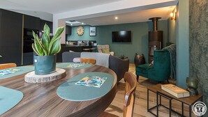 A modern living room with a wooden dining table, a sofa, and a green armchair. There is a kitchen area in the background with a clock and a television. - Chalet le René-Sens - BOUTX (COULEDOUX)