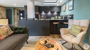 A modern living room with a sofa, armchair, and coffee table, a kitchen with dark cabinets and a bar area, and a skylight above the kitchen. - Chalet le René-Sens - BOUTX (COULEDOUX)