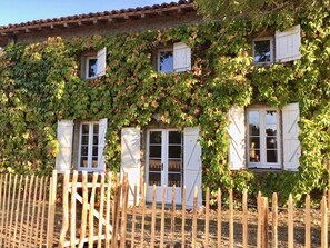 A rustic building with a stone facade, wooden shutters on windows, and a wooden fence in front. - La Maison des Champs (CINTEGABELLE)