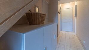 Un couloir avec un sol carrelé de carreaux blancs, une porte blanche au fond et un panier posé sur un meuble.