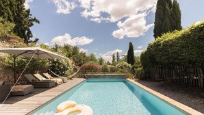 Una piscina rectangular con muelle de madera y sillas de descanso, rodeada de valla de madera y vegetación.