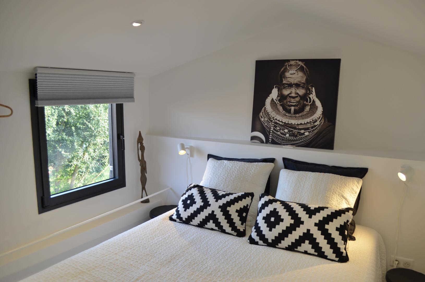 Un dormitorio con una cama, almohadones estampados, una ventana con persianas, una lámpara de pared y un cuadro enmarcado.