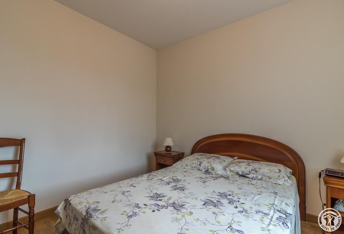 Un dormitorio con una cama individual, una mesita de noche de madera con una lámpara, una silla y una pequeña mesa con un libro.