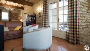 A cozy living room with a sofa, armchairs, and a table. There is a window with curtains, a fireplace, and a stone wall. - Domaine de Blanconne (Léguevin)