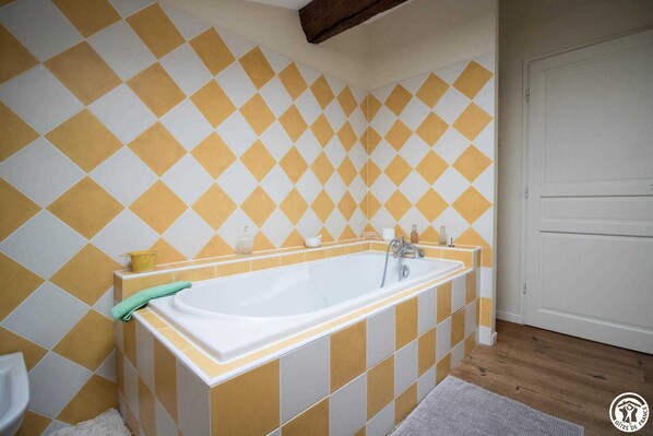 Ein Badezimmer mit einer großen Wanne, gelb-weißen Karokacheln, Holzdielen und einer weißen Tür.