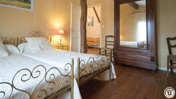 A bedroom with a bed, a nightstand, a lamp, a chair, a mirror, and a door leading to another room. - Domaine de Blanconne (Léguevin)