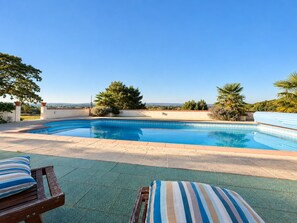 Ein Schwimmbecken vor einem wolkenlosen, blauen Himmel, umgeben von Grünpflanzen und mit Blick auf die Landschaft.