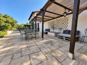 Un patio exterior con suelo de piedra, mobiliario de metal y una zona de estar cubierta.