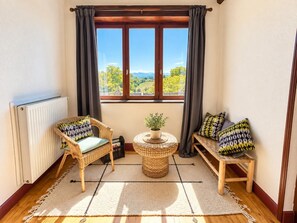 Una habitación acogedora con vistas a un paisaje verde, una silla de mimbre, una mesita redonda con una planta y un banco con cojines estampados.